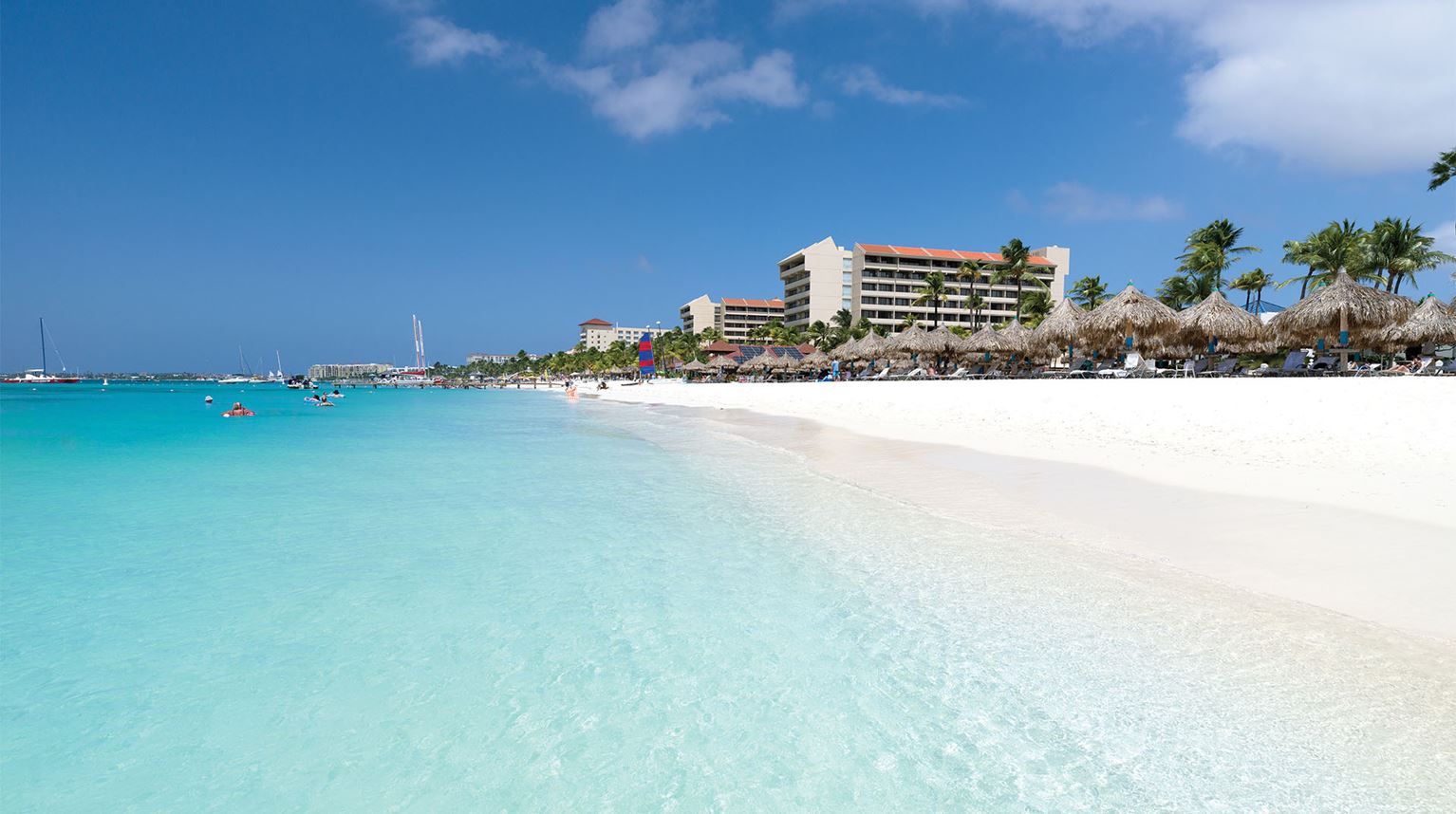 White sandy beach and turquoise water with palm trees and a mid-rise building in the background.