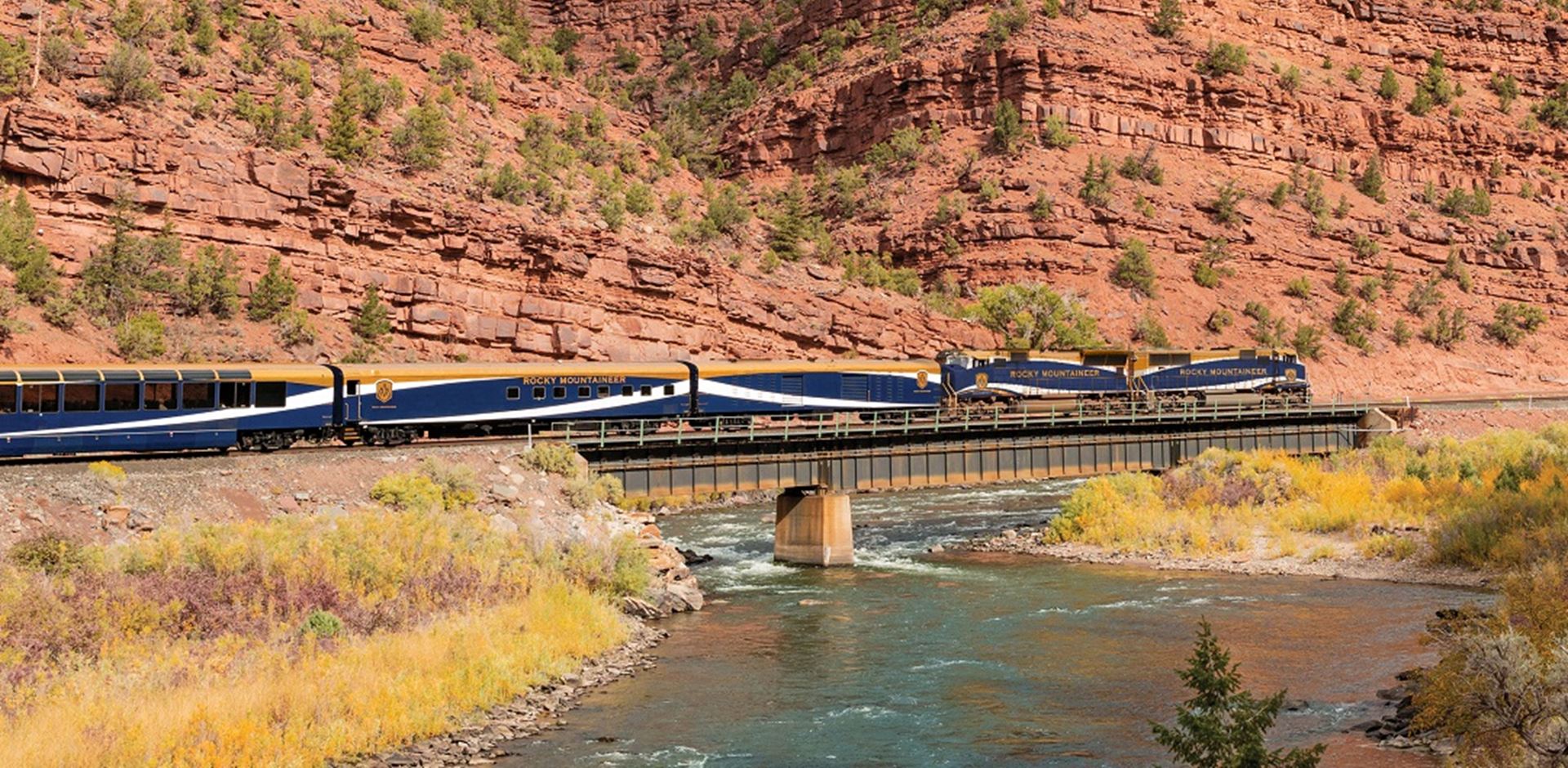 A blue Rocky Mountaineer train against red rock mountains 