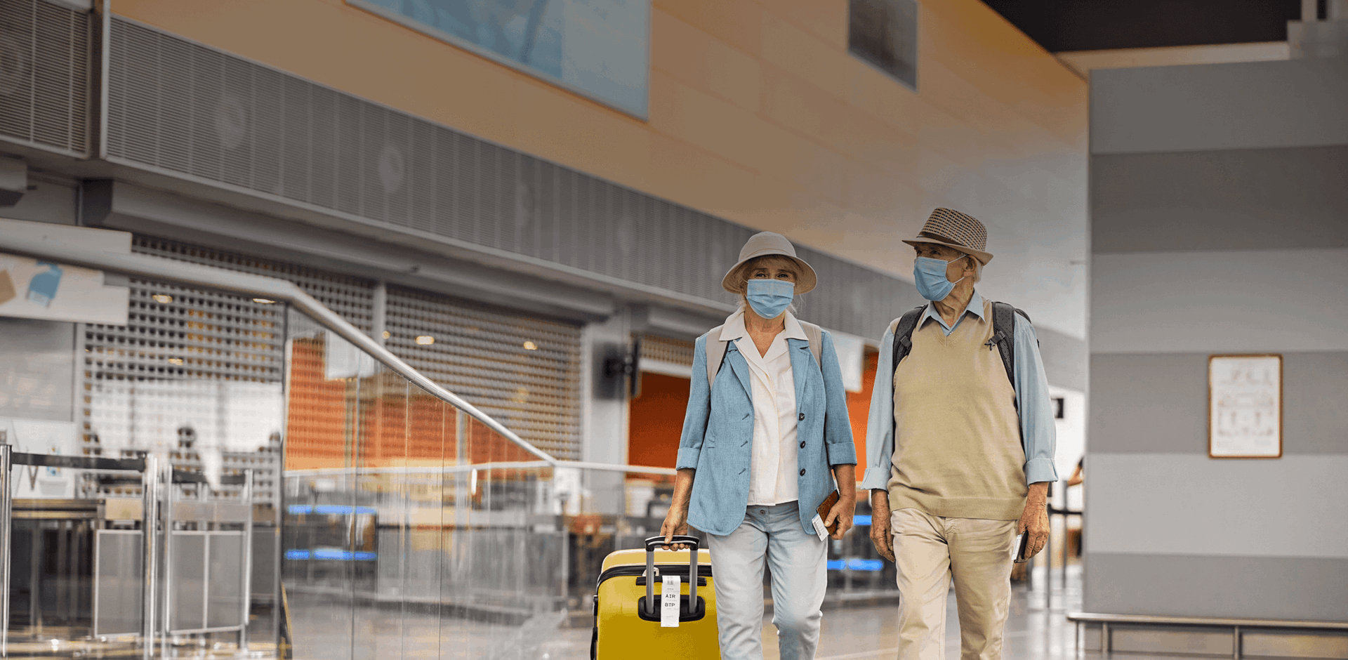 Aged tourist couple with suitcases walking along the airport corridor in masks.