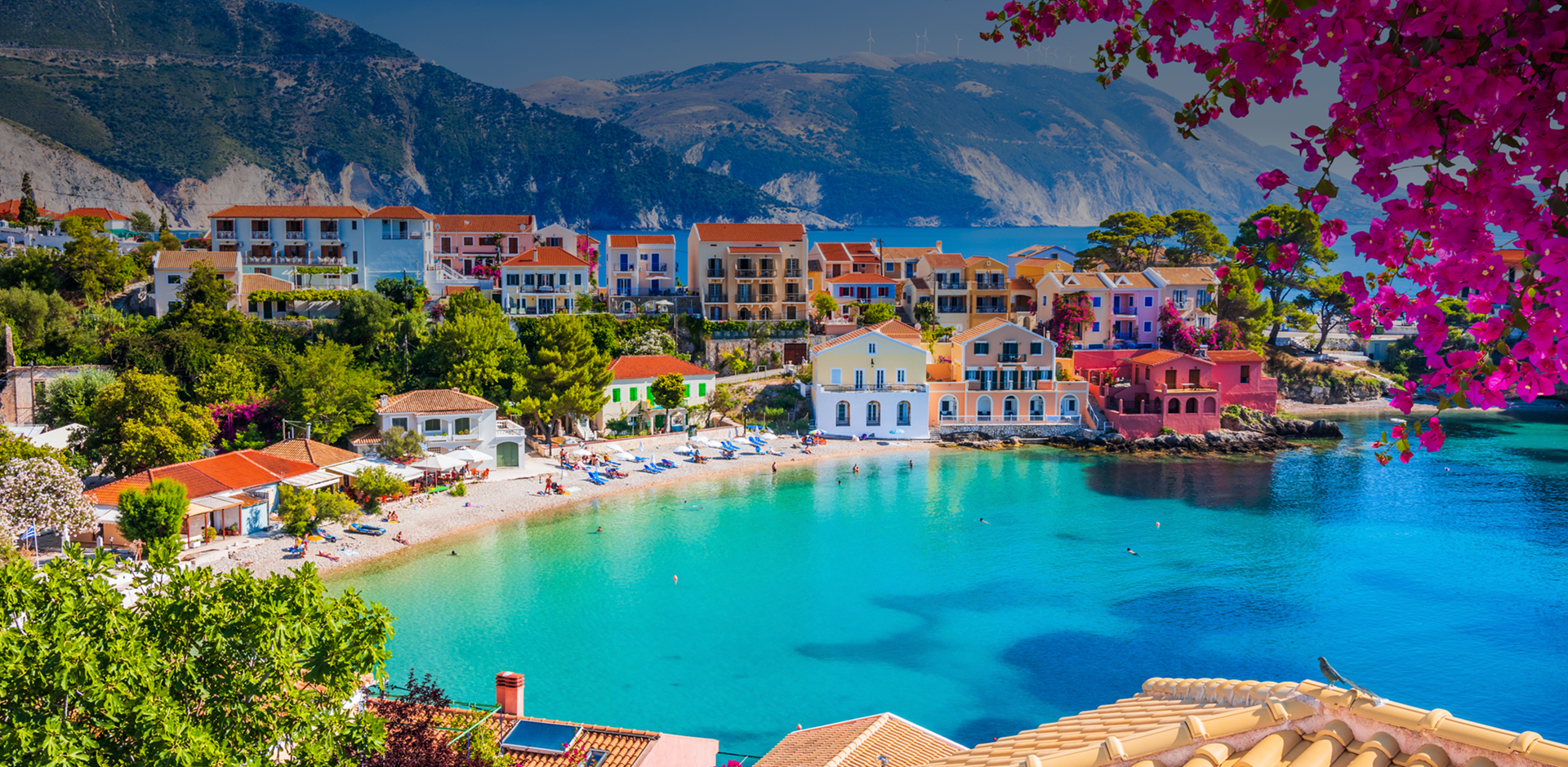 View of colourful houses with hills behind them, small beach with clear water in front and a tree with blooming pink flowers overhanding from the top of the image, Village of Assos in Kefalonia, Greece.