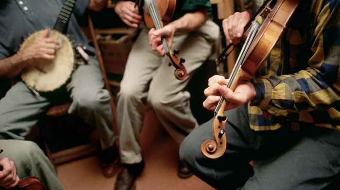 Four Bluegrass musicians sitting in circle on chairs and rehearsing on different instruments.